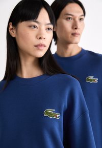 Sudadera azul hecha de una tela suave, que presenta un escote redondo y un logotipo de cocodrilo verde bordado en el pecho.