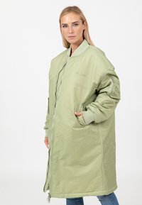 Vrouw in een oversized lichtgroene bomberjack met ritssluiting, ribbelmanchetten en kraag, staand met één hand in de pocket tegen een witte achtergrond.