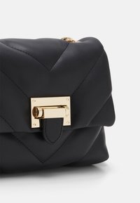 Černá prošívaná kožená kabelka se zlatým obdélníkovým zapínáním na otočnou klapku a řetízkovým popruhem částečně viditelným na bílém pozadí.