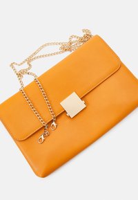 Oranžová kožená clutch taška se zlatým čtvercovým zapínáním a odnímatelným zlatým řetízkovým ramínkem na bílém pozadí.