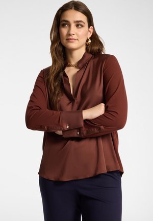 Femme aux longs cheveux bruns portant un chemisier marron à manches longues et un pantalon foncé, les bras croisés, regardant sur le côté sur un fond blanc.
