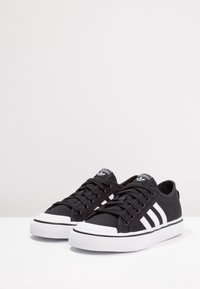 Black canvas sneakers with white rubber sole and toe cap. Features three white stripes on the side and round laces. Classic low-top design.