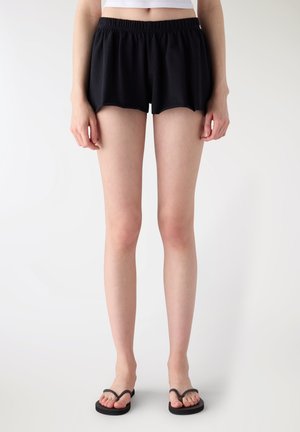 Donna con pantaloncini neri a vita elastica e infradito nere, in piedi davanti a uno sfondo bianco uniforme.