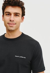 Czarna bawełniana koszulka z krótkim rękawem, okrągłym dekoltem i białym haftowanym logo na lewej piersi. Gładka tekstura i klasyczny krój.