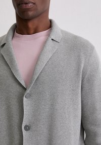 Hellgraue Strickjacke aus strukturiertem Stoff, mit Reverskragen und zwei dunklen Knöpfen. Über einem blassrosa Rundhals-Pullover getragen.