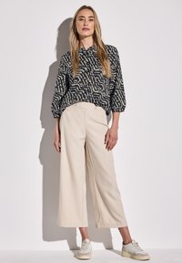 Blusa nera e beige con motivo, dotata di colletto arricciato, abbinata a pantaloni larghi color avorio. La modella indossa sneakers chiare.