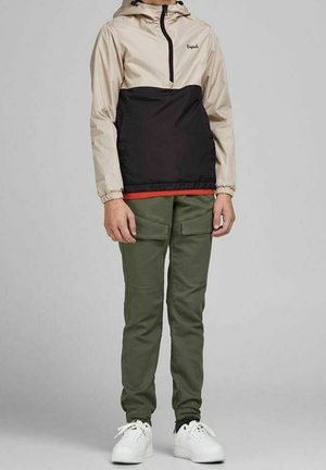 Kind trägt beige-schwarze Kapuzenjacke, olivgrüne Cargohose und weiße Turnschuhe, steht vor einfachem grauem Hintergrund.