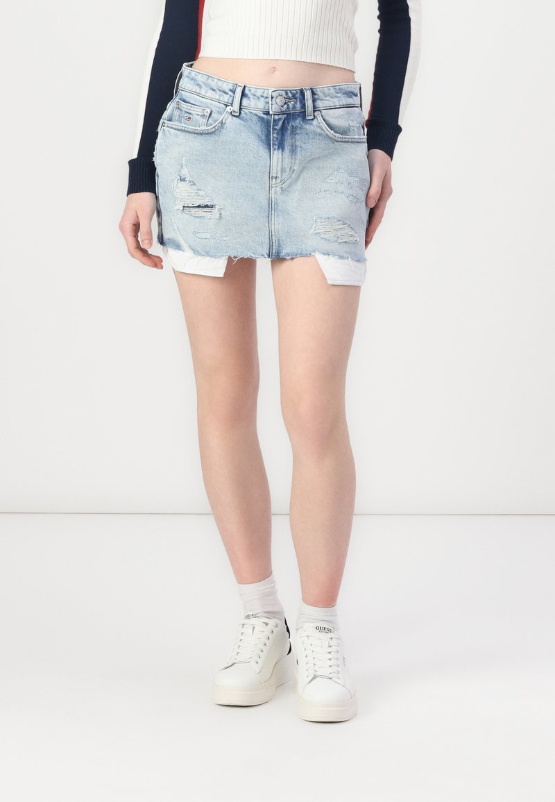 Denim mini szoknya, világoskék mosású, kopottas szélű, öt zsebes dizájnnal, fehér sportcipővel és szürke zoknival párosítva.
