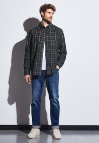 Tmavě kostkovaná košile na knoflíky v zelené, modré a hnědé barvě, kombinovaná s modrými džíny a světlými teniskami, umístěná na čistém bílém pozadí.