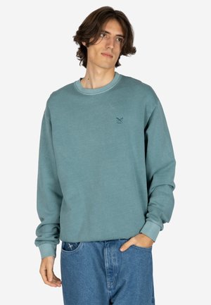 Junger Mann mit mittellangen Haaren, der einen türkisen Langarmpullover und blaue Jeans trägt, steht mit einer Hand in der Tasche.