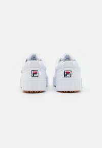 Sneakers bianchi in materiale sintetico con suola spessa, caratterizzati da un piccolo logo in blu navy e rosso sul tallone. Suola in gomma testurizzata per una maggiore aderenza.