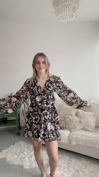 Μαύρο και άσπρο φόρεμα τύπου wrap με λουλουδάτο μοτίβο και μακριά διάφανα μανίκια, με βολάν στο τελείωμα. Το μοντέλο στέκεται πάνω σε υφασμένο χαλί, χαμογελώντας.