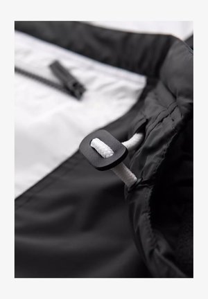 Schwarz-weißer Jackenstoff, der einen Nahaufnahme eines schwarzen Kordelzuges mit einem weißen Seil zeigt, mit einer seitlichen Reißverschlusstasche.