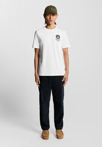 Hvid kortærmet t-shirt med en marineblå grafik på venstre bryst, parret med mørke bukser og en grøn kasket med et gult logo.