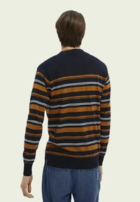 Man som bär en marinblå, brun och vit randig tröja och blå byxor, sedd bakifrån mot en enfärgad bakgrund.