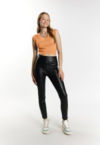 Orange ribbet crop top, højtaljede sorte læderbukser og hvide sneakers med mintgrønne accenter. Modellen står med den ene hånd på hoften.
