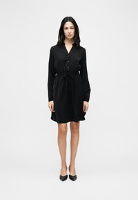 Γυναίκα που στέκεται φορώντας μαύρο φόρεμα μήκους μέχρι τα γόνατα με μακριά μανίκια, κουμπωτό μπροστινό μέρος, ζώνη με κορδόνι στη μέση και μαύρες μυτερές γόβες, σε λευκό φόντο.