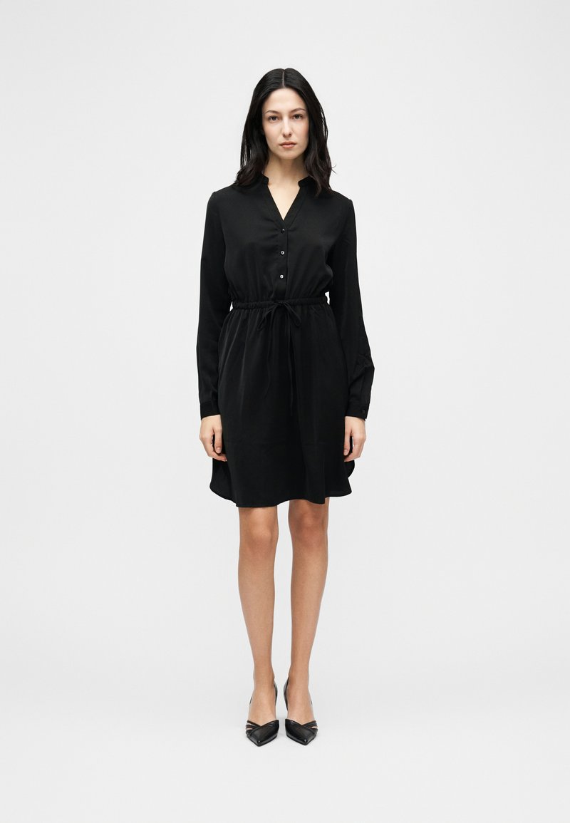 Γυναίκα που στέκεται φορώντας μαύρο φόρεμα μήκους μέχρι τα γόνατα με μακριά μανίκια, κουμπωτό μπροστινό μέρος, ζώνη με κορδόνι στη μέση και μαύρες μυτερές γόβες, σε λευκό φόντο.