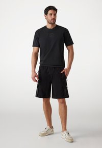 Schwarzes T-Shirt mit kurzen Ärmeln und dezentem Logo, kombiniert mit schwarzen Cargo-Shorts mit Seitentaschen und Kordelzug, ergänzt durch weiße Sneakers.