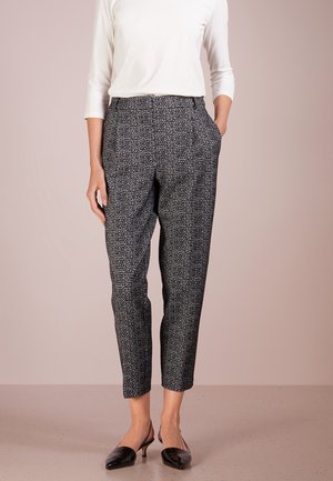 Vrouw draagt een zwart-witte, hoog getailleerde broek tot op de enkels met patroon, een wit topje met lange mouwen en zwarte lage hakken.