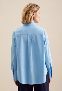 Camicia azzurro chiaro a maniche lunghe con bottoni, caratterizzata da una piega dietro, colletto a punta e maniche con polsini. Tessuto liscio con una silhouette aderente.