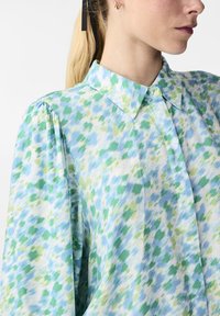 Blusa leggera con colletto in tonalità di blu e verde con un motivo maculato. Presenta maniche lunghe con dettagli arricciati.