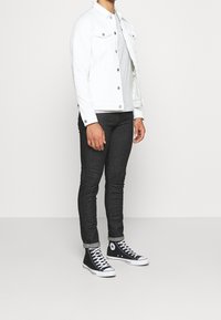 Chaqueta de mezclilla blanca, camiseta gris, vaqueros ajustados oscuros y zapatillas altas negras. La chaqueta presenta bolsillos frontales y botones plateados.
