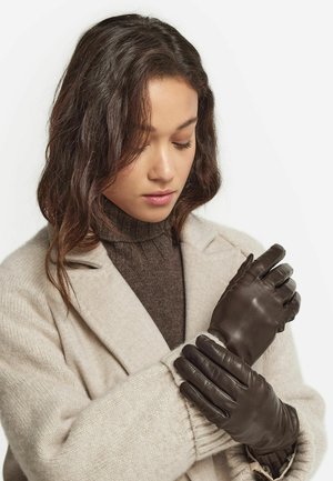 Kvinde med langt mørkt hår iført en beige frakke og en brun rullekrave trøje, der tager mørkebrune læderhandsker på foran en ensfarvet baggrund.