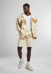 Beige hoodie with large brown text, paired with matching shorts. White t-shirt underneath, beige sneakers, and subtle texture throughout.