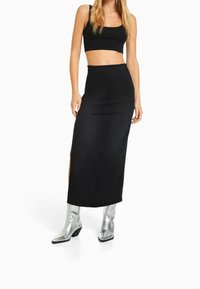 Falda midi negra de talle alto con abertura lateral, hecha de tejido suave. La modelo lleva un top corto a juego y botas de tobillo metálicas plateadas.