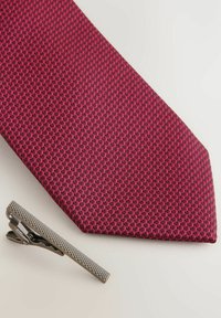 Struktureret bordeaux slips med et geometrisk mønster og en sølv slipsenål med en ribbet overflade, placeret ved siden af slipset.