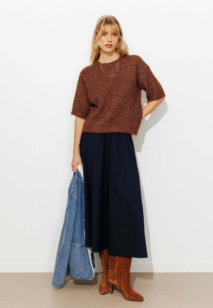 Femme portant un pull marron à manches courtes, une longue jupe bleu marine et des bottes en daim marron, tenant une veste en jean bleue dans sa main gauche.