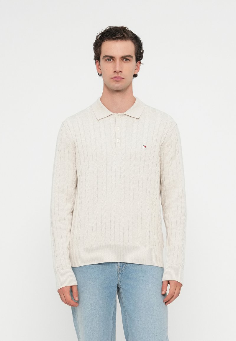 Piqué-Pullover in Creme mit Kabelmuster, geripptem Kragen und Bündchen, mit einem kleinen Logopatch auf der linken Brust. Getragen mit blauen Jeans.