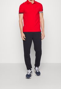 Rød polo med korte ærmer, marineblå sweatpants med "HILFIGER" tekst og marineblå sneakers med hvide detaljer. Ensfarvet baggrund.