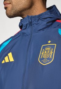 Tmavomodrá bunda s vysokým golierom, obsahujúca červené a svetlomodré akcenty, žlté logo Adidas a emblém národného tímu Španielska s korunou.