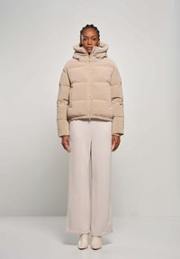 Vrouw in een beige gewatteerde jas met capuchon en lichte, wijde broek, staand tegen een effen lichtgrijze achtergrond.
