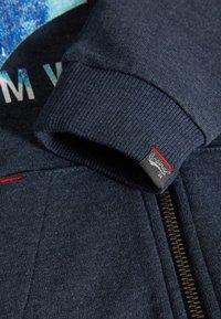 Mörkblå sweatshirttyg med ribbad mudd och strukturerad design. Har en synlig dragkedja och en märkesetikett med röda accenter.