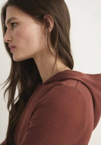 Smeđa dukserica s kapuljačom koja se ističe mekanom teksturom, uskim krojem i glatkim završetkom, koju nosi osoba s dugom tamnom kosom.