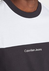 Camiseta de manga corta con un diseño en bloques de color blanco y gris oscuro. Presenta un escote redondo y el logo de "Calvin Klein Jeans" en blanco.