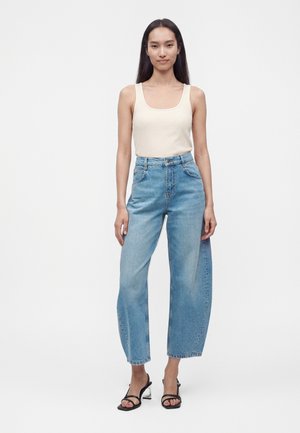 Donna in piedi che indossa una canotta bianca senza maniche, jeans celesti a vita alta e sandali neri con cinturini e tacco su sfondo bianco.