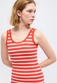 Νεαρή γυναίκα με αμάνικο τοπ με κόκκινες και άσπρες ρίγες, φοράει ασημένια σκουλαρίκια κρεμαστά και κοιτάζει προς τα δεξιά της, με φόντο ένα απλό ανοιχτόχρωμο φόντο.