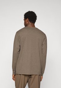 Hombre con cabello corto y rizado, vistiendo una camisa de manga larga marrón y pantalones marrones, de pie mirando hacia atrás contra un fondo claro y liso.