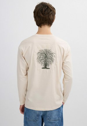 Hombre que lleva una camisa de manga larga color crema con un gráfico de palma verde y texto circular descolorido en la parte trasera, combinado con jeans de mezclilla azul.