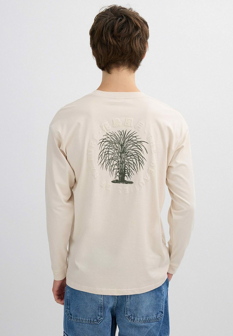 Hombre que lleva una camisa de manga larga color crema con un gráfico de palma verde y texto circular descolorido en la parte trasera, combinado con jeans de mezclilla azul.