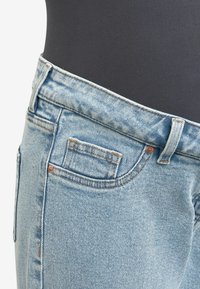 Jeans in denim azzurro chiaro con vita alta, tasche inclinate e cuciture marroni a contrasto. Il tessuto ha una texture leggermente sbiadita.