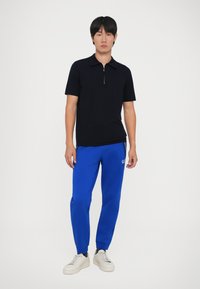 Crna polo majica kratkih rukava s zip ovratnikom, u skladu s plavim trenerkama s bočnim džepovima i logotipom, upotpunjeno bijelim tenisicama.