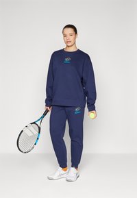 Marineblå sweatshirt og joggebukser lavet af blødt stof, med et logo på brystet og låret, parret med hvide sneakers og et tennisketsj.