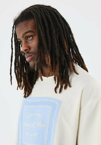 Camiseta de algodón de corte grande en color beige con un gráfico azul y un borde blanco intrincado junto con texto. El modelo tiene dreadlocks largas, oscuras y de tonos múltiples.
