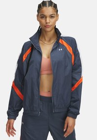 Navy windbreaker with orange and white diagonal stripes, ribbed cuffs, and a zip closure. Lightweight fabric, casual fit.