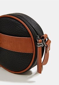 Pyöreä musta nahkalaukku, jossa on ruskea nahkakoriste ja kohokuvioitu logo. Laukussa on vetoketjullinen suljin ja irrotettava olkahihna. Pintamateriaali on teksturoitu.